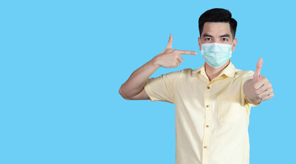 COVID-19 Coronavirus portrait handsome young asian man wearing yellow shirt and mask protection from covid 19 and thumbs up sign isolated on blue background in studio. Asian people. COVID-19 concept.