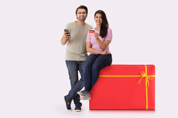 A young woman sitting on a gift shaped stool with a man beside,showing credit card and phone.