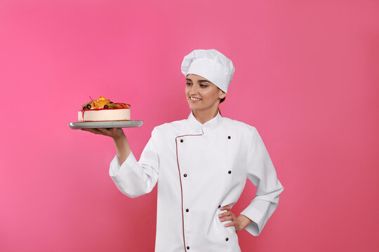 Happy professional confectioner in uniform holding delicious cake on pink background