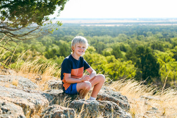 Naklejka na ściany i meble Young boy sitting on rock on hilltop smiling with sweeping landscape view in background