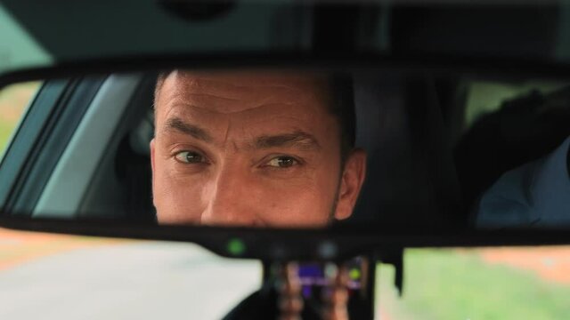 Close-up eyes of a young man are reflected in the rear-view mirror of a car. The guy looks at the road, then at the camera. The concept of travel and recreation by car on vacation or weekend. UHD 4K.