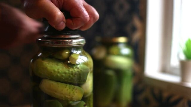 A mans hand rolls up a jar of cucumbers preserves. A person prepares pickles in the kitchen using a seamer