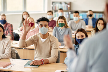 Black student with face mask raises his hand to ask question during lecture at college classroom.