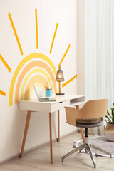 Laptop, stationery and lamp on white table near wall with painted sun indoors. Interior design