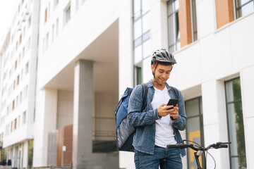 Medium shot of cheerful handsome male courier with thermo backpack standing with bicycle in city street and using navigation app on phone. Delivery man looking for client address looking smartphone.
