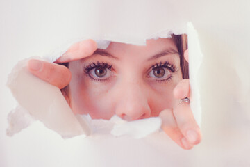 Brunette woman in isolation looking through a hole in white paper with uncertainty