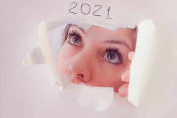 Brunette woman looking through a hole in white paper that has 2021 handwritten sign on it with surprise and uncertainty 
