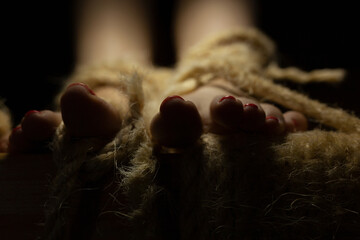 Female feet with red nail polish tied to the wooden beam with shabby rope, in the dark