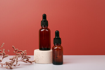 Selective focus, two dark transparent glass bottles with skin serum or oil for manicure on beige gypsum podium against dark red background decorated with dry flower. Skin care concept with copy space
