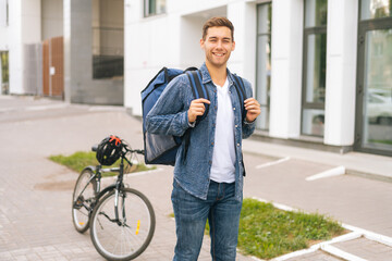 Fototapeta premium Portrait of smiling handsome young delivery man with thermo backpack standing on city street, on background of bicycle and office building, looking at camera.