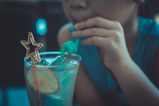 Close up and selective focus shot of lemonade soda in cocktail glass with fancy straw and lemon slice decoration in blurred background of adorable kid girl drinking for her refreshment.