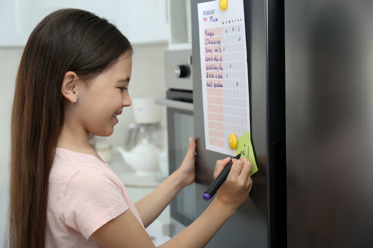 Little girl drawing funny face on note near to do list in kitchen