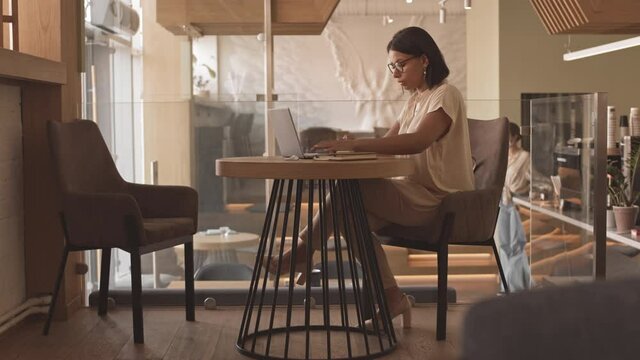 Wide side-view of elegant Mixed-Race woman sitting at table, using portable computer in modern restaurant