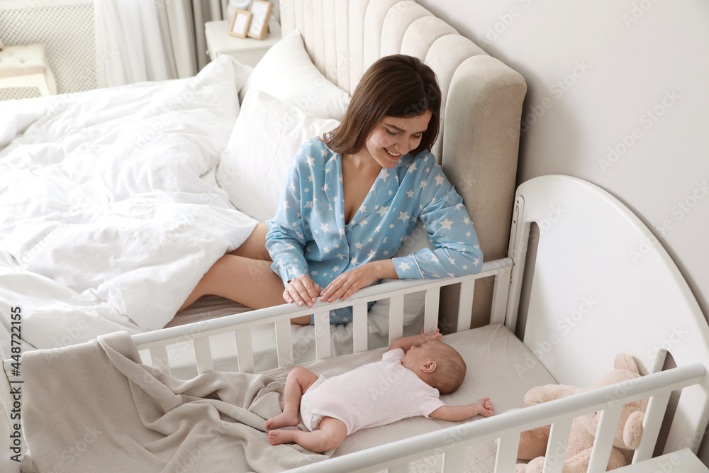 Poster happy young mother near crib with sleeping newborn baby in bedroom