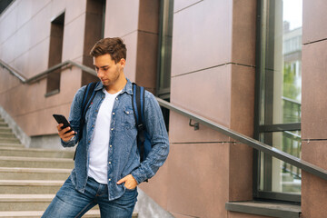 Medium shot of handsome delivery man with large thermo backpack holding phone, looking at screen, using navigation app. Courier male getting order standing on stairs of office building.