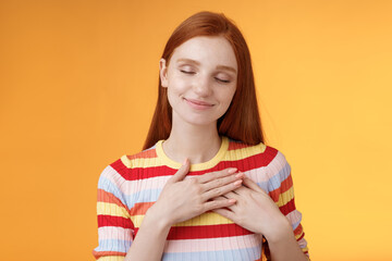 Heart keeps warm save dearest memories inside. Smiling tender heartwarming redhead lovely girl touch chest grinning closed eyes recall lovely nice moment standing romantic orange background