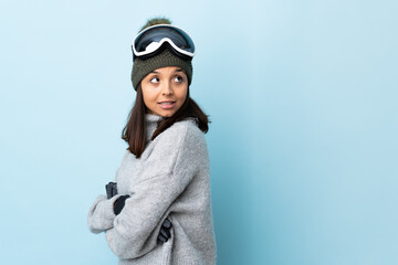 Mixed race skier girl with snowboarding glasses over isolated blue background with arms crossed and happy.