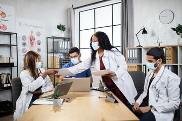 Obraz premium Two men and two women in lab coats with stethoscopes sitting at table and using modern gadgets, afro lady giving papers for colleagues. Multiracial doctors analyzing diagnosis of severe patient.