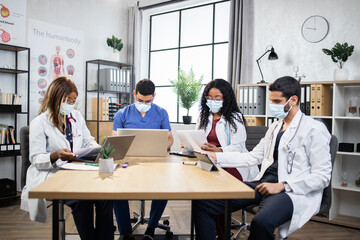 Fototapeta premium Group of four multiracial doctors in masks using modern gadgets while examining patient's record during consilium. International colleagues sitting at room discussing various way of treatment.