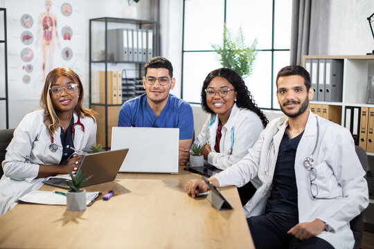 Two Arab men and two Afro-American women scientists doctors, sitting at table and using modern gadgets. Multiracials doctors working together on new scientific project, smiling at camera