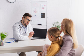 Family doctor pointing at laptop near mother and daughter in clinic