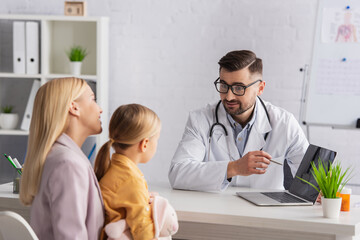 Fototapeta premium Blurred mother and girl looking at doctor in eyeglasses pointing at laptop