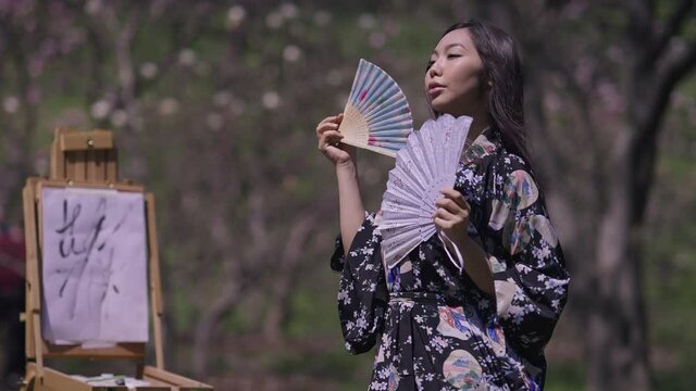 Gorgeous Asian young woman in kimono dancing with fans in spring sakura garden smiling as blurred easel with canvas painting at background. Portrait of confident Japanese lady outdoors on sunny day
