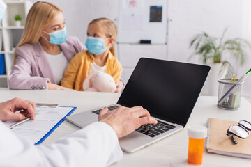 Doctor using laptop near pills and mother with daughter in medical masks on blurred background