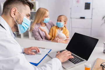 Pediatrician in medical mask using laptop near pills and family in clinic