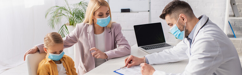 Mother in protective mask pointing at kid near pediatrician writing on clipboard, banner
