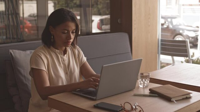 Medium close-up of young Mixed-race woman sitting at table in restaurant, typing email on portable computer, smiling
