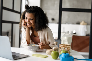Black young woman working with laptop while having breakfast at home