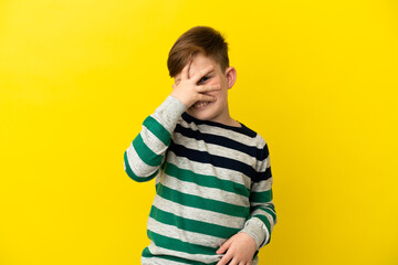 Little redhead boy isolated on yellow background covering eyes by hands and smiling
