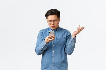 Frustrated and confused asian guy in glasses looking puzzled at mobile phone screen, raising hand...