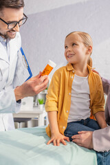 Positive doctor with clipboard holding pills near smiling girl on medical couch and woman