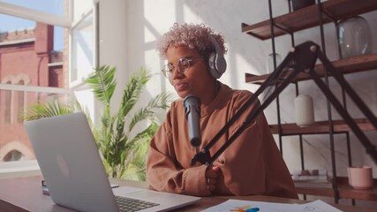 African American female wearing wireless headphones greets his followers and talking on microphone, working at communication webinar in living room. Black woman blogger social media putting on blog. - Powered by Adobe