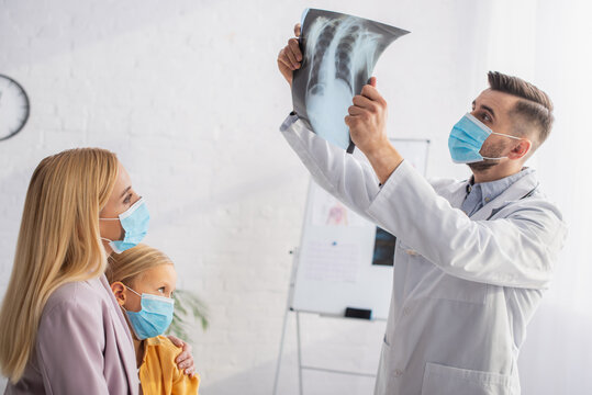 Pediatrician in medical mask looking at fluorography near mother and patient