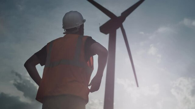 A male engineer or repair worker stands next to a wind turbine. Man looks at the propeller and fan blades. White safety construction helmet. Alternative energy sources. Green Fuel Generator