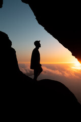 Chico en cueva con vistas al atardecer, mar de nubes.