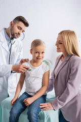 Obraz premium Smiling kid looking at camera near mom and doctor with stethoscope