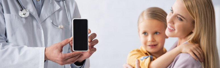 Doctor holding smartphone with blank screen near smiling mother and kid, banner