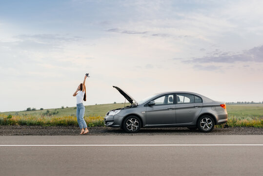 A young girl stands near a broken-down car in the middle of the highway during sunset and tries to call for help on the phone. Breakdown and repair of the car. Waiting for help.