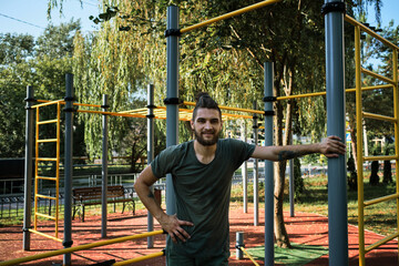 Fototapeta premium Active lifestyle concept. Young man is standing on equipped outdoor exercise area and resting. Banner with male fitness trainer. Replenishes reserve of strength and energy before strength training.