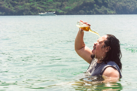 A Cool Drunk Asian Guy With Long Hair Pours A Bottle Of Beer On His Head While Standing Chest Deep Waters. Unwinding At The Beach.