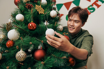 handsome man decorating christmas tree with baubles and looking at camera at hom