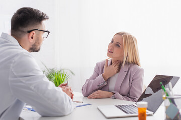 woman touching sore throat during consultation with doctor near laptop