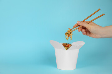 Woman eating wok noodles with chopsticks from box on light blue background, closeup. Space for text