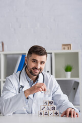 happy doctor looking at camera while making pyramid with medical symbols on desk