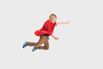 Portrait of little blond boy, pupil flying like super hero in red cape isolated on white studio background. Copyspace for ad. Childhood, education, emotion concept.