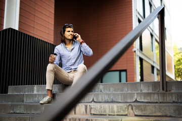 Young man sitting on the stairs drinking coffee. Handsome businessman using the phone while drinking coffee